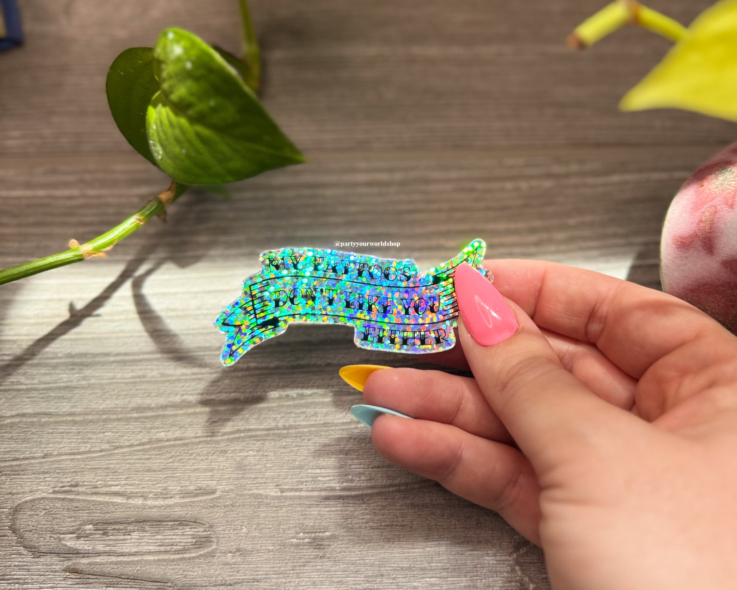 Hand holding a colorful, glittery sticker "my tattoos don't like you either" on a wooden surface