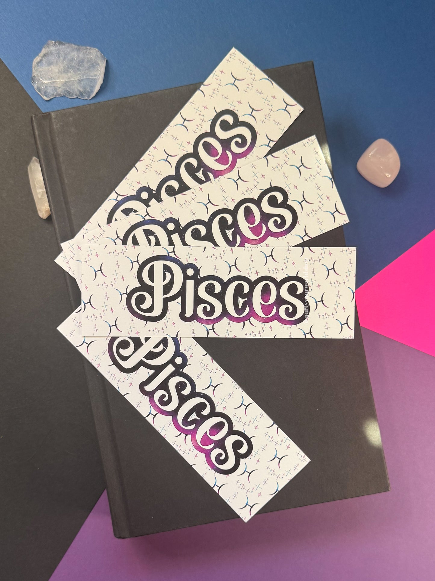 Three rectangular bookmarks with the word 'Pisces' in a stylized black and pink gradient font, arranged on top of a dark gray book against a blue and purple background with small crystal props.