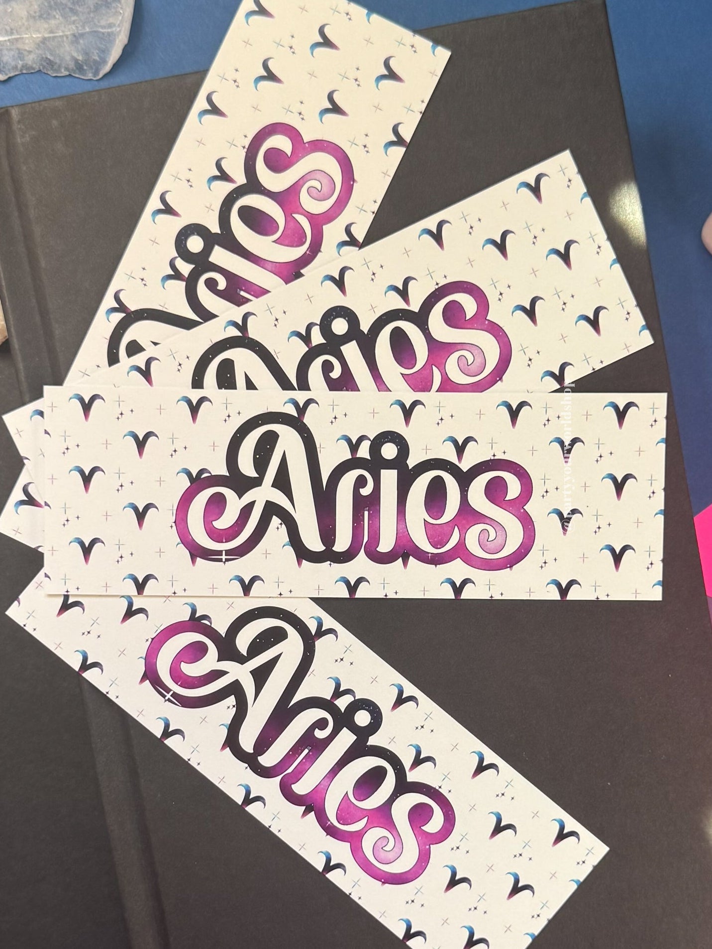 Three bookmarks with the word 'Aries' in stylized pink, purple, and black script on a white background with small black bird motifs, arranged on top of a dark book against a blue and purple surface with crystal props.