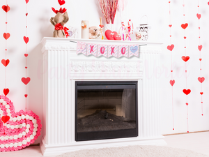  A styled mantelpiece decorated for Valentine’s Day with the candy heart banner spelling "XOXO." The background includes heart garlands, teddy bears, and other pink and red Valentine’s-themed decorations.