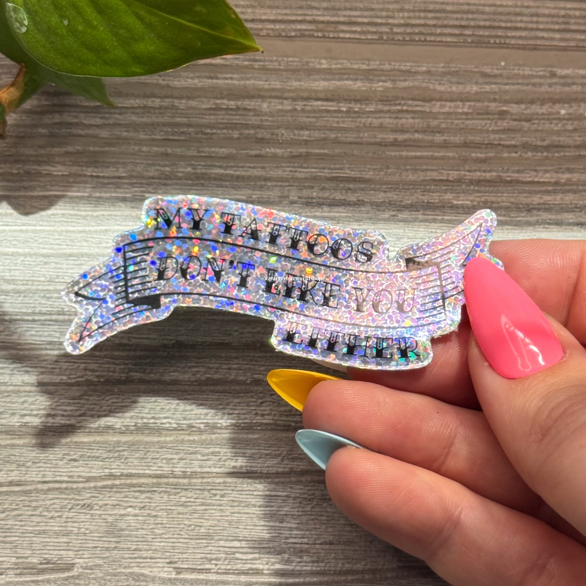 Hand holding a colorful, glittery sticker "my tattoos don't like you either" on a wooden surface