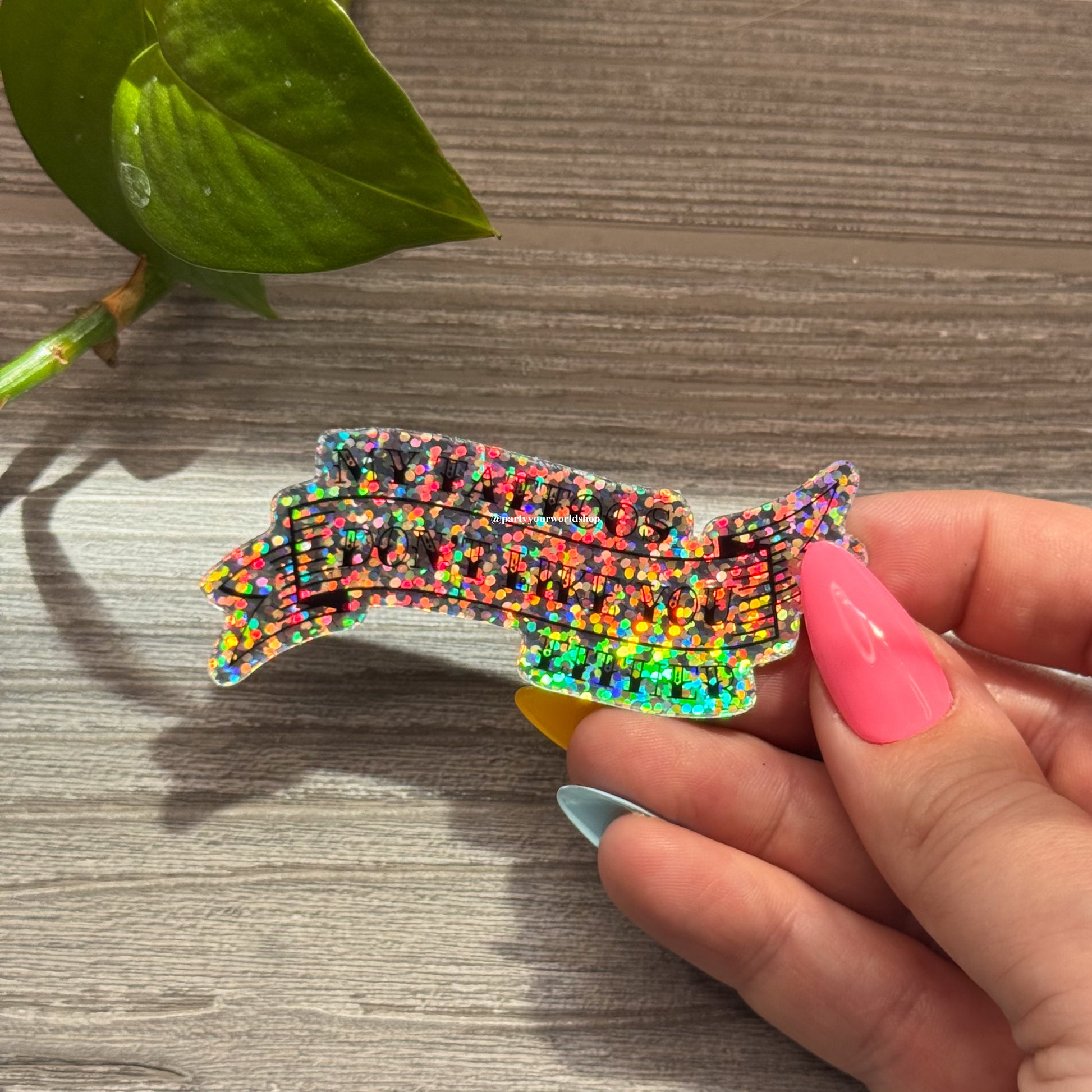 Hand holding a colorful, glittery sticker "my tattoos don't like you either" on a wooden surface