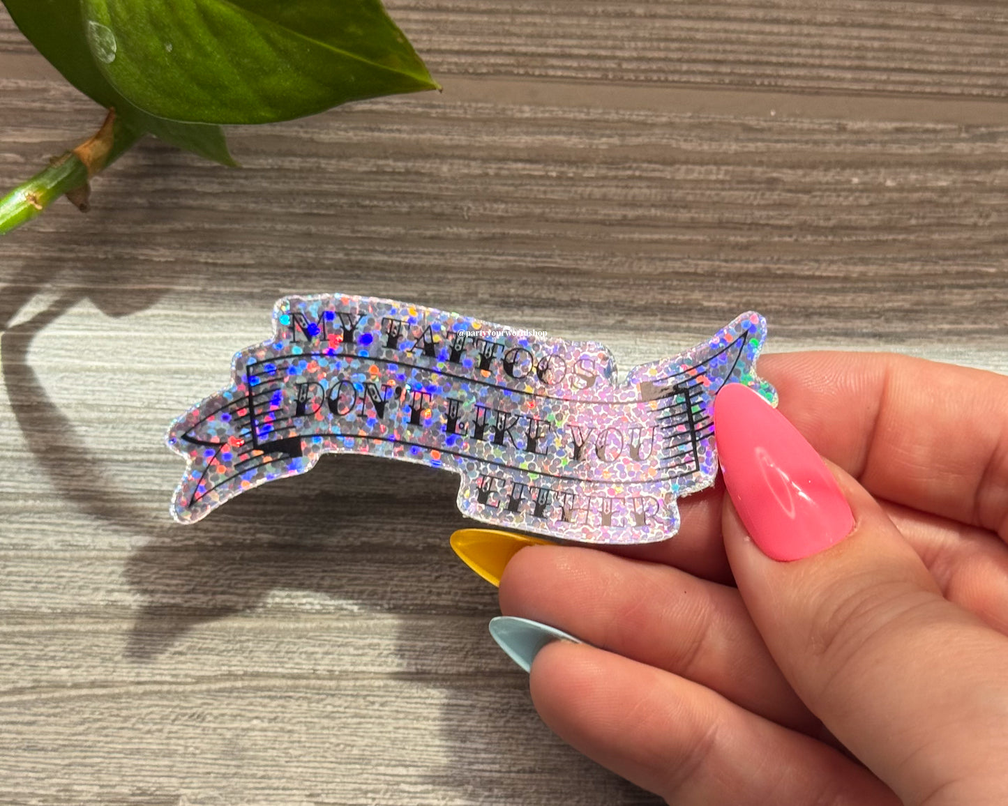 Hand holding a colorful, glittery sticker "my tattoos don't like you either" on a wooden surface