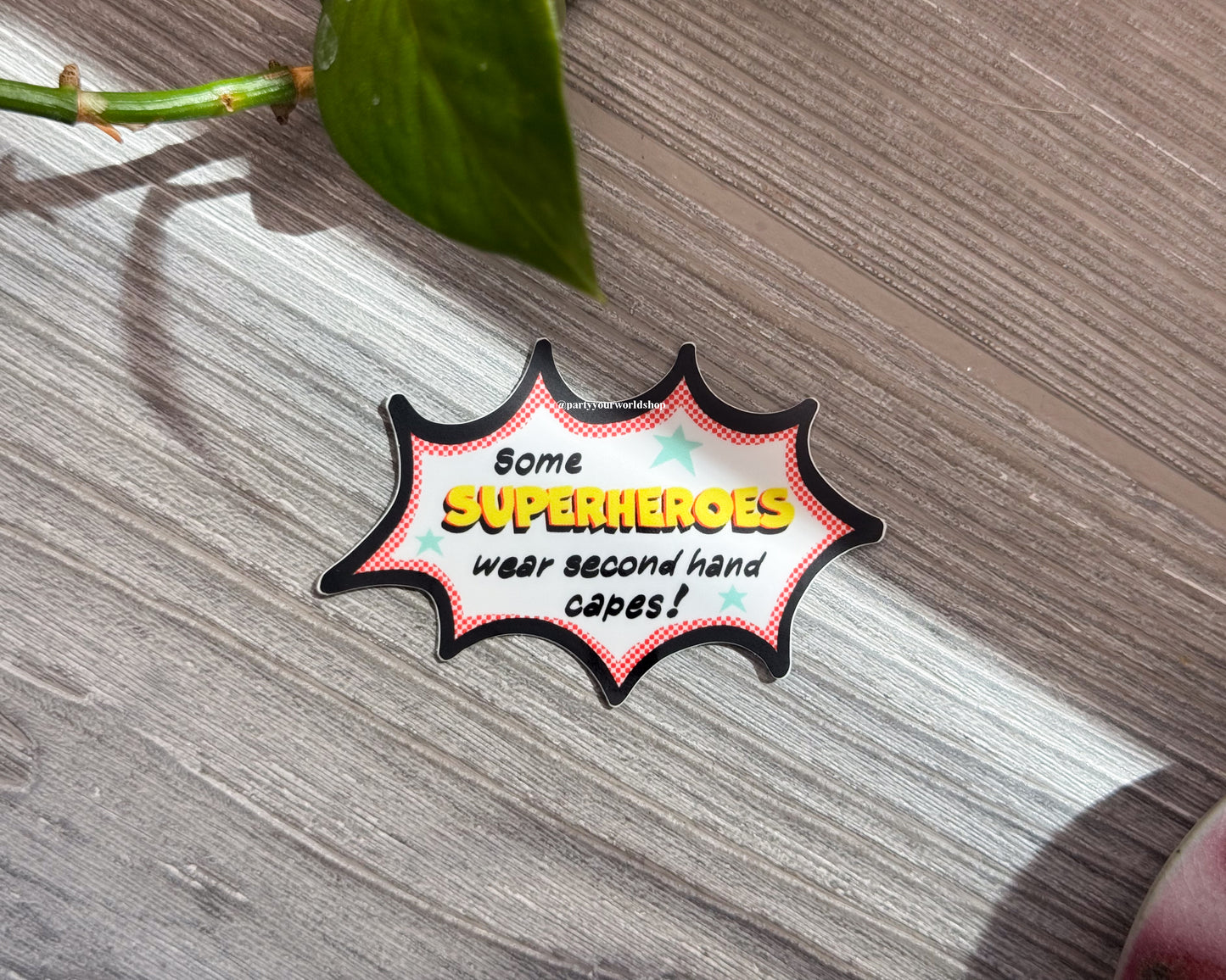 Superhero-themed "some superheroes wear second hand capes" sticker with text against wood background