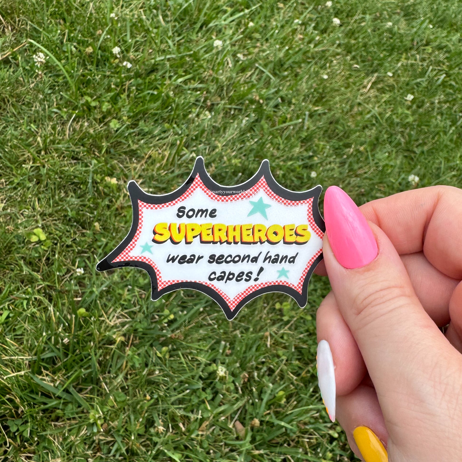 Hand holding a superhero-themed "some superheroes wear second hand capes" sticker with text against a grass background