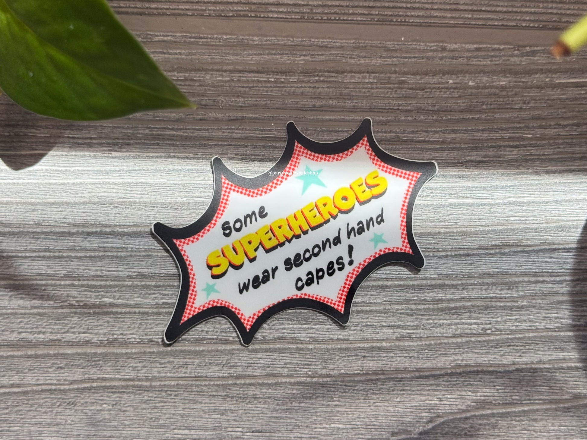Comic-style badge with text 'Some superheroes wear second hand capes!' on a wooden surface.