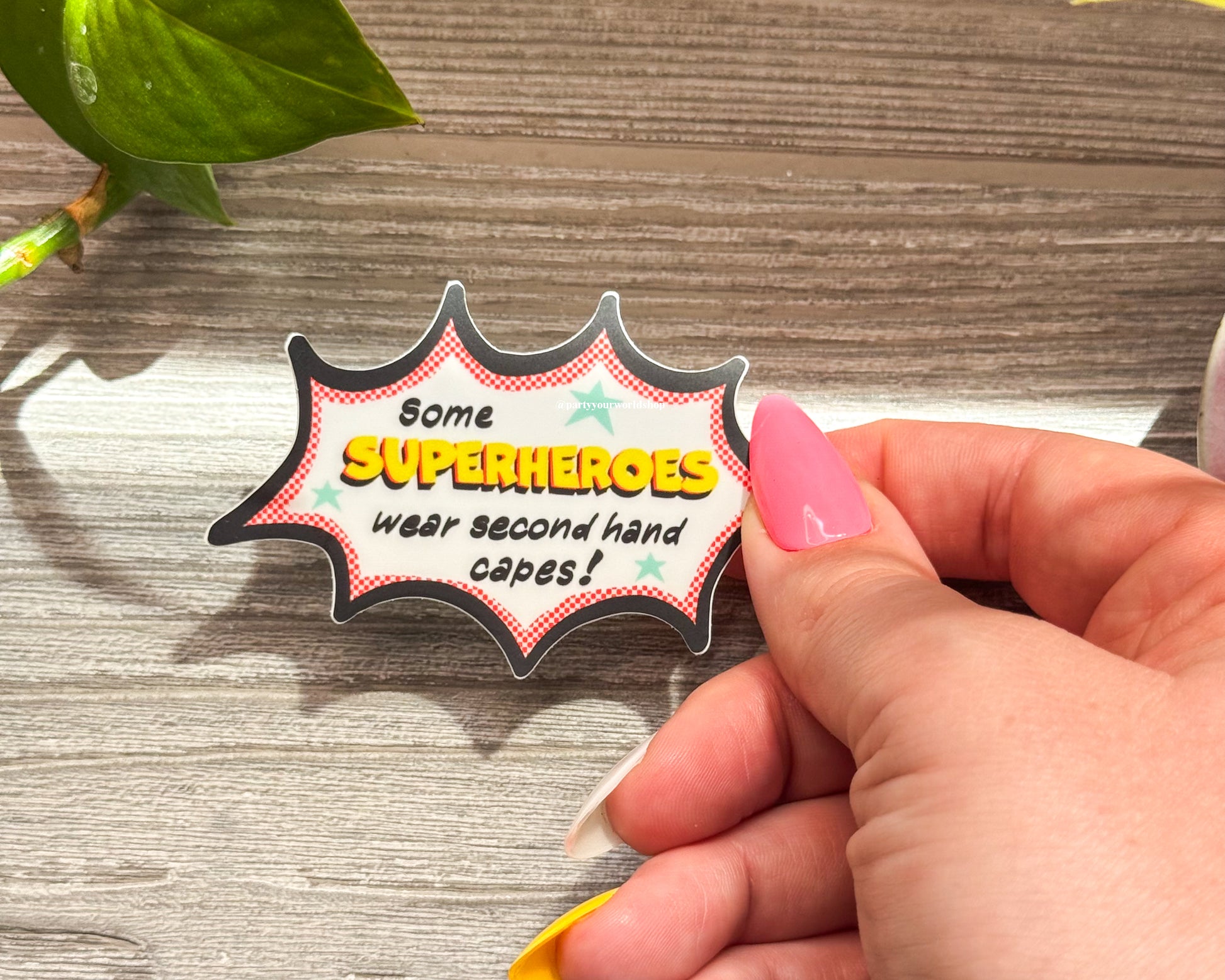 Hand holding a superhero-themed sticker with text on a wooden surface