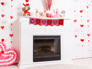 Close-up of a Valentine’s Day banner hanging on a white fireplace mantle, surrounded by heart-shaped decorations and Valentine's Day-themed items.