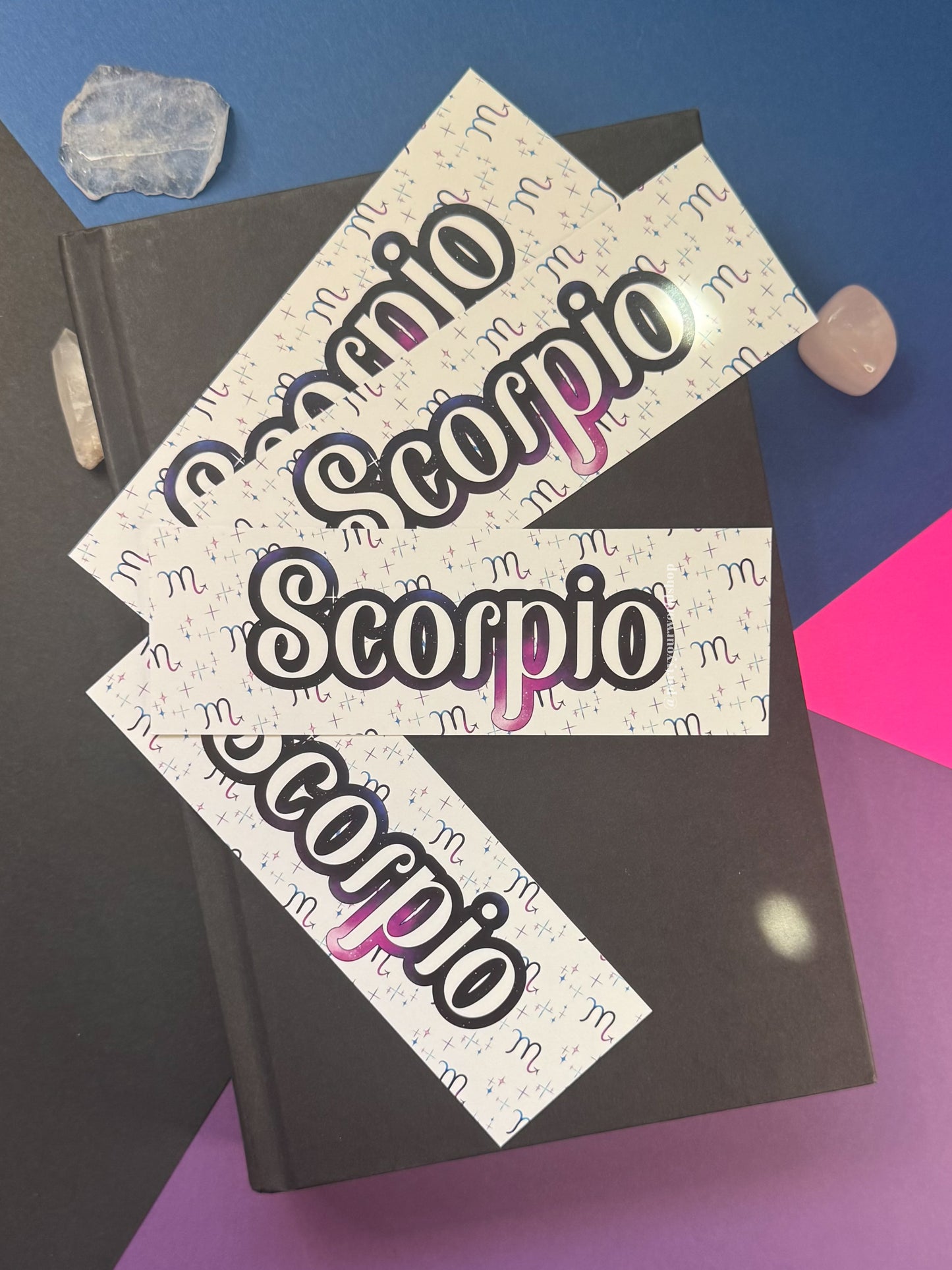 Three bookmarks with the word 'Scorpio' in large black outlined lettering and a pink gradient accent, arranged on top of a dark book with crystal stones around it.