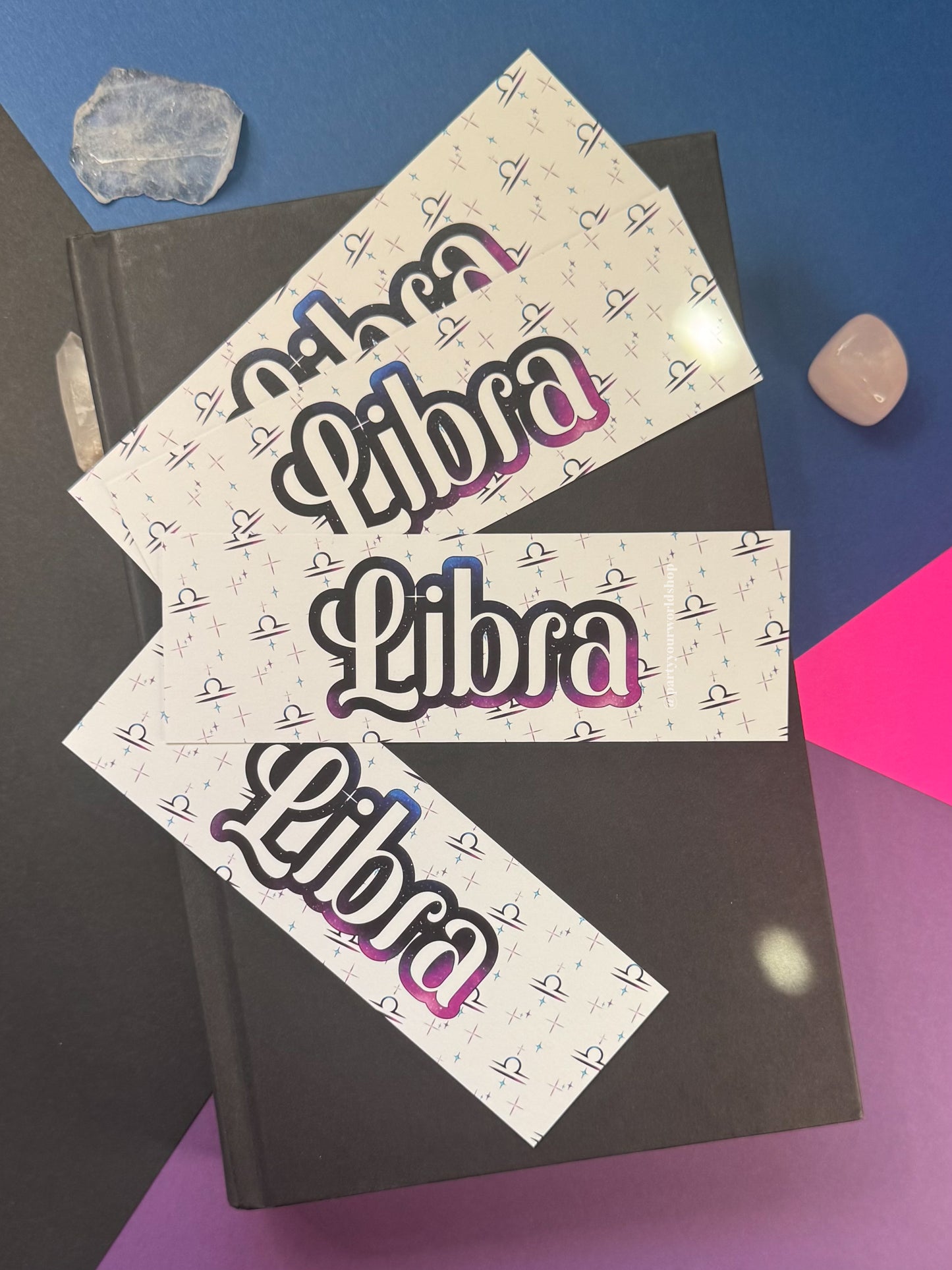 Three bookmarks with the word 'Libra' in a stylized black, pink, and blue gradient font, decorated with a small repeating cross pattern. The labels are arranged on top of a black notebook, with crystal stones on a blue and purple background.