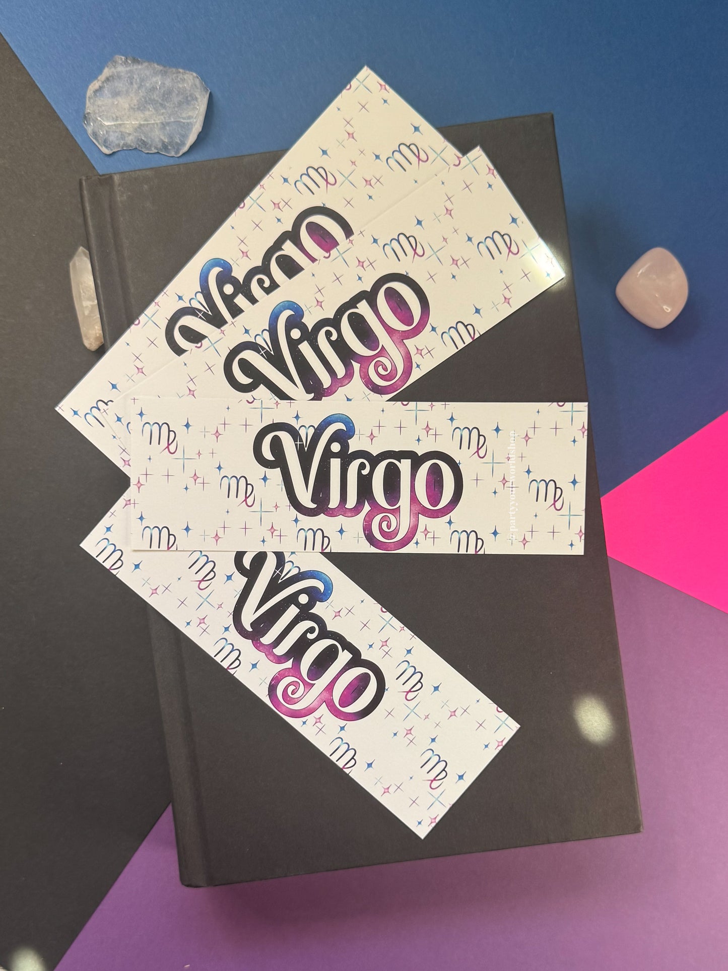 Three bookmarks with the word 'Virgo' in large black script outlined in pink and blue, arranged on top of a dark book against a blue, purple, and pink background with small crystal stones.