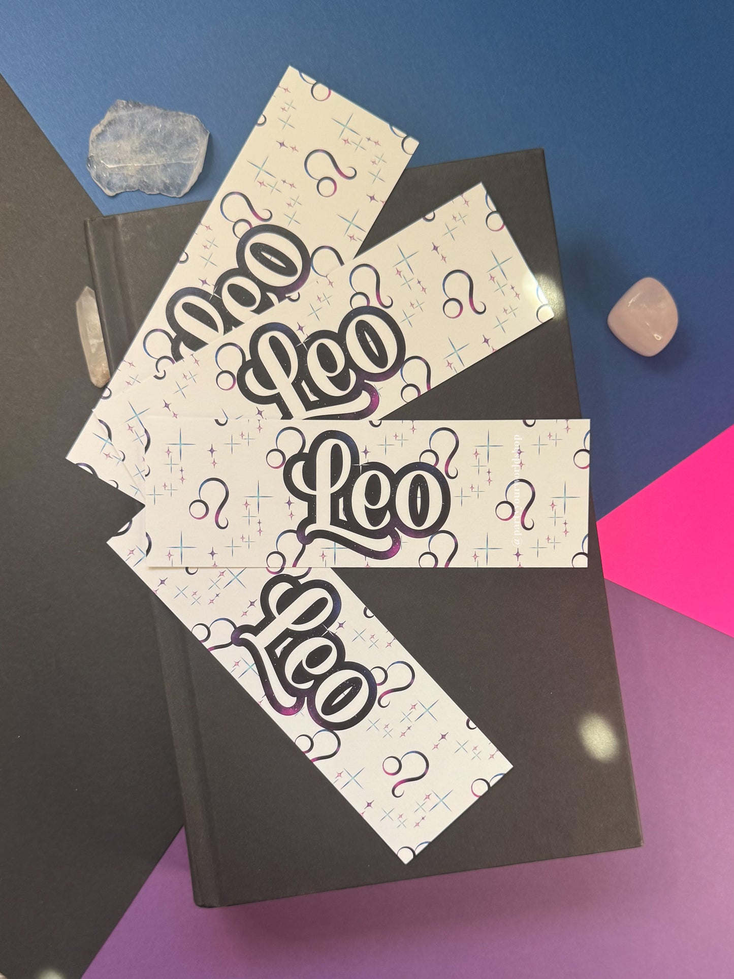 Three white paper bookmarks printed with the word 'Leo' in a stylized black and purple font, arranged on top of a black hardcover book. The scene is styled with crystals and bright blue, pink, and purple background panels.