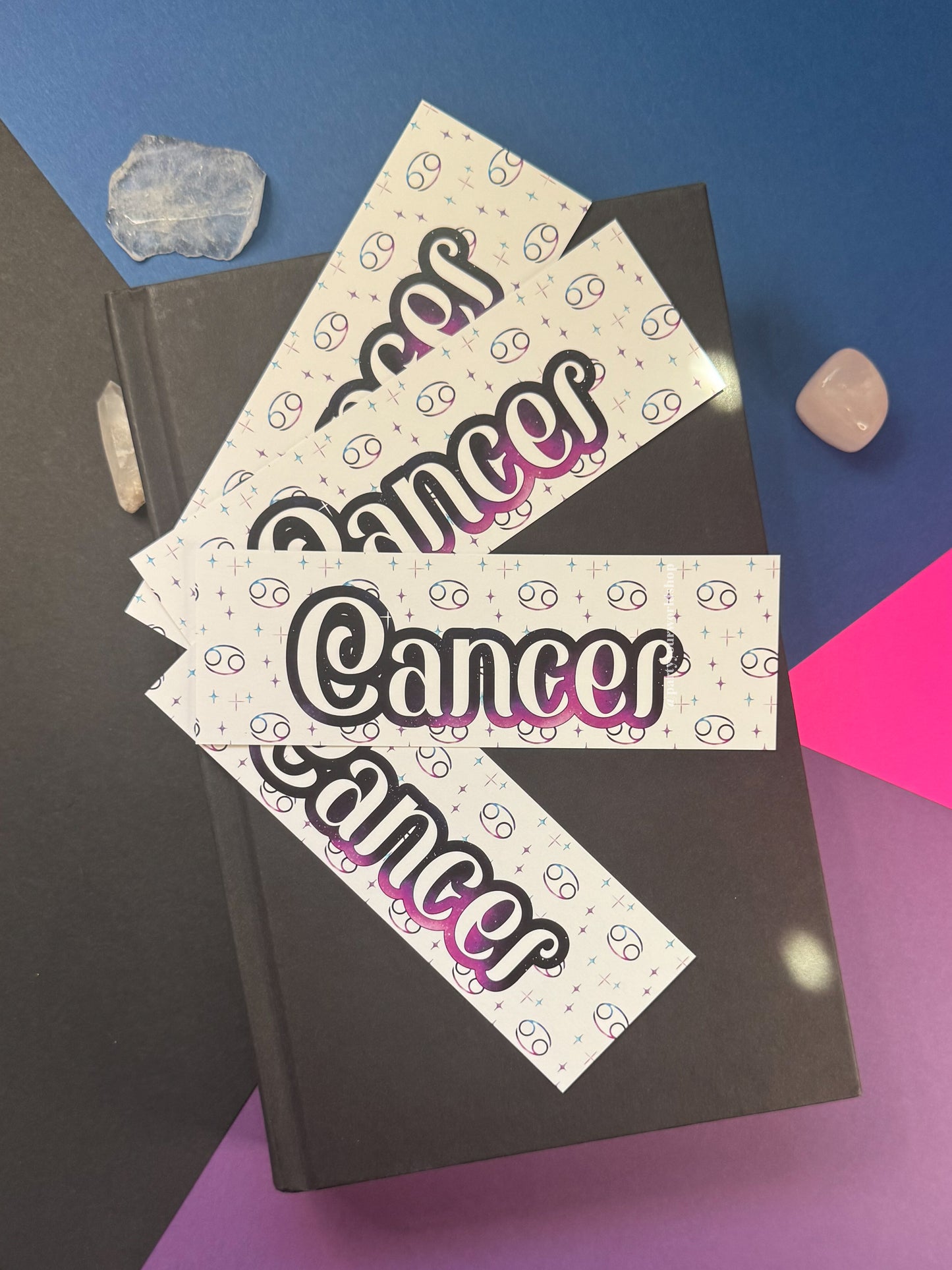 Three rectangular  bookmarks with the word "Cancer" in large black script and a pink-to-purple gradient outline, arranged on top of a dark hardcover book. The cards have a white background with small black doodle-like symbols.