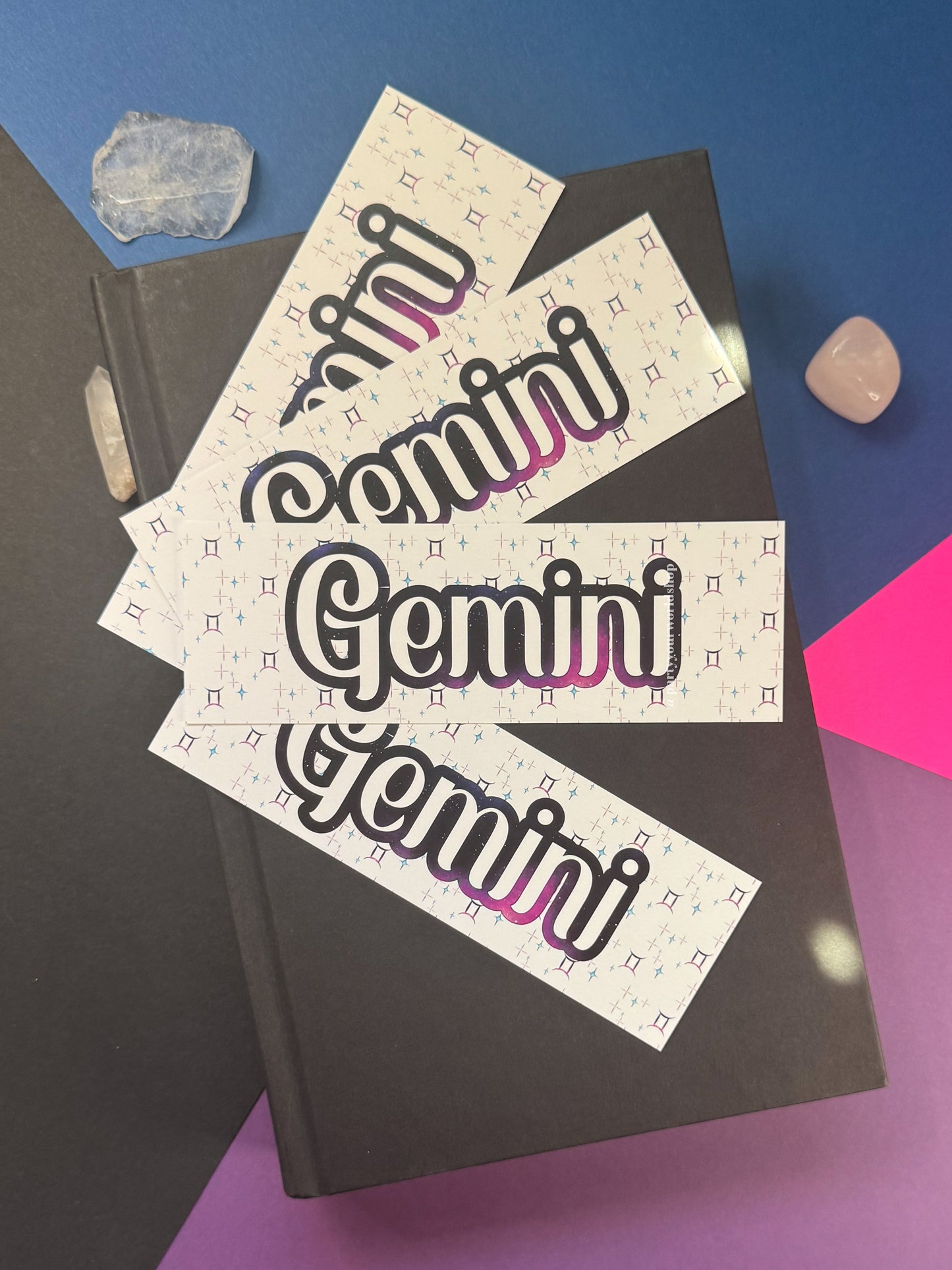 Three white rectangular bookmarks 
 with the word 'Gemini' in large black outlined lettering and a pink-to-purple gradient accent, arranged on top of a dark notebook against a blue and purple background with crystals.