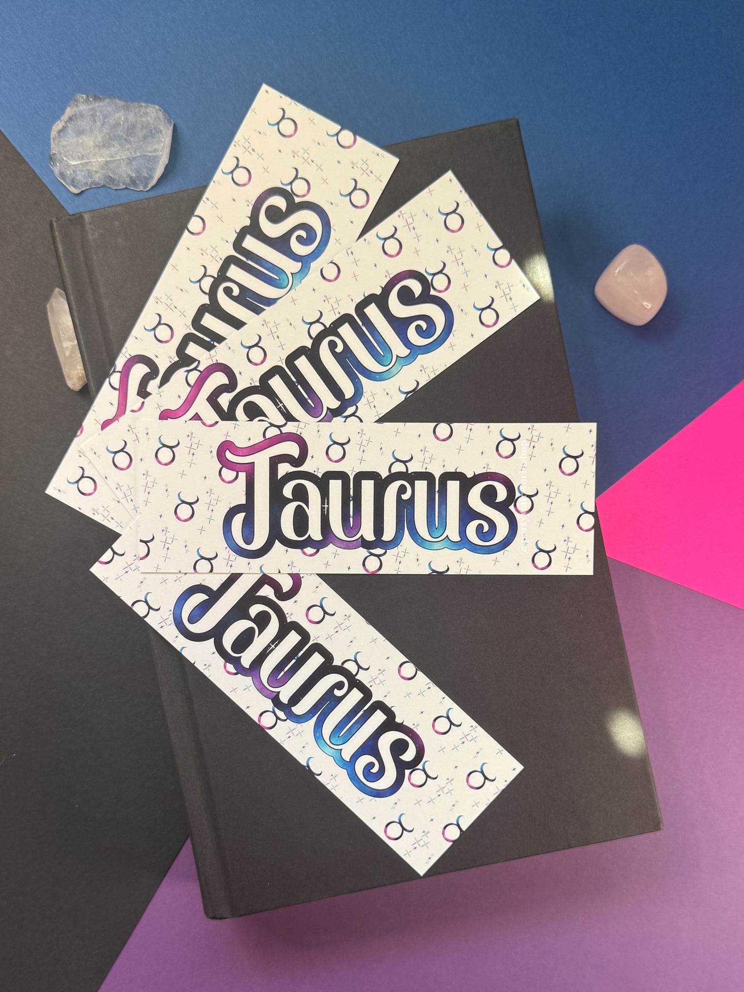 Three rectangular bookmarks printed with the word "Taurus" in large stylized lettering and a repeating zodiac symbol pattern, arranged on top of a black book against a blue, purple, and pink background with crystal props.