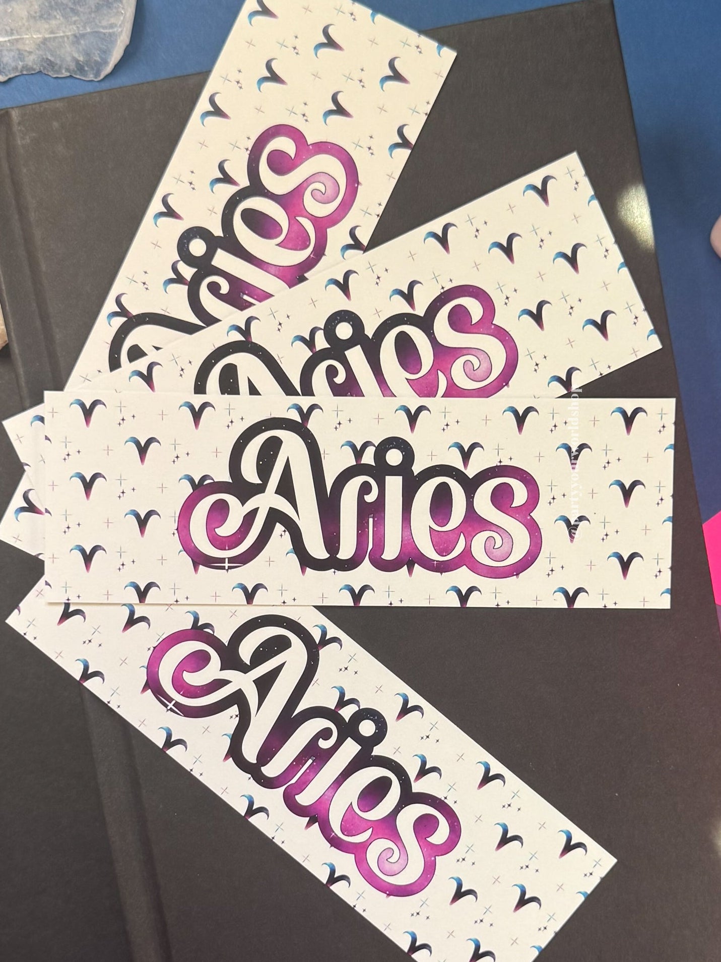 Three bookmarks with the word 'Aries' in stylized pink, purple, and black script on a white background with small black bird motifs, arranged on top of a dark book against a blue and purple surface with crystal props.