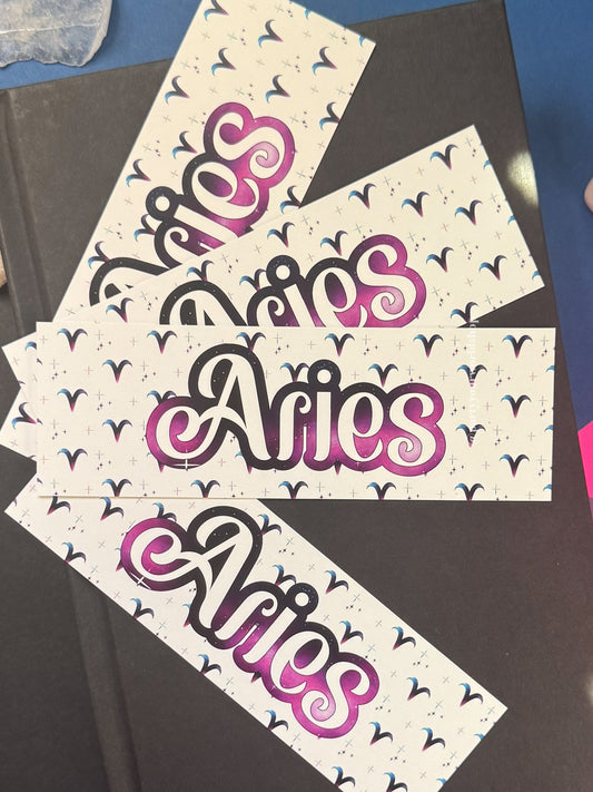 Three bookmarks with the word 'Aries' in stylized pink, purple, and black script on a white background with small black bird motifs, arranged on top of a dark book against a blue and purple surface with crystal props.