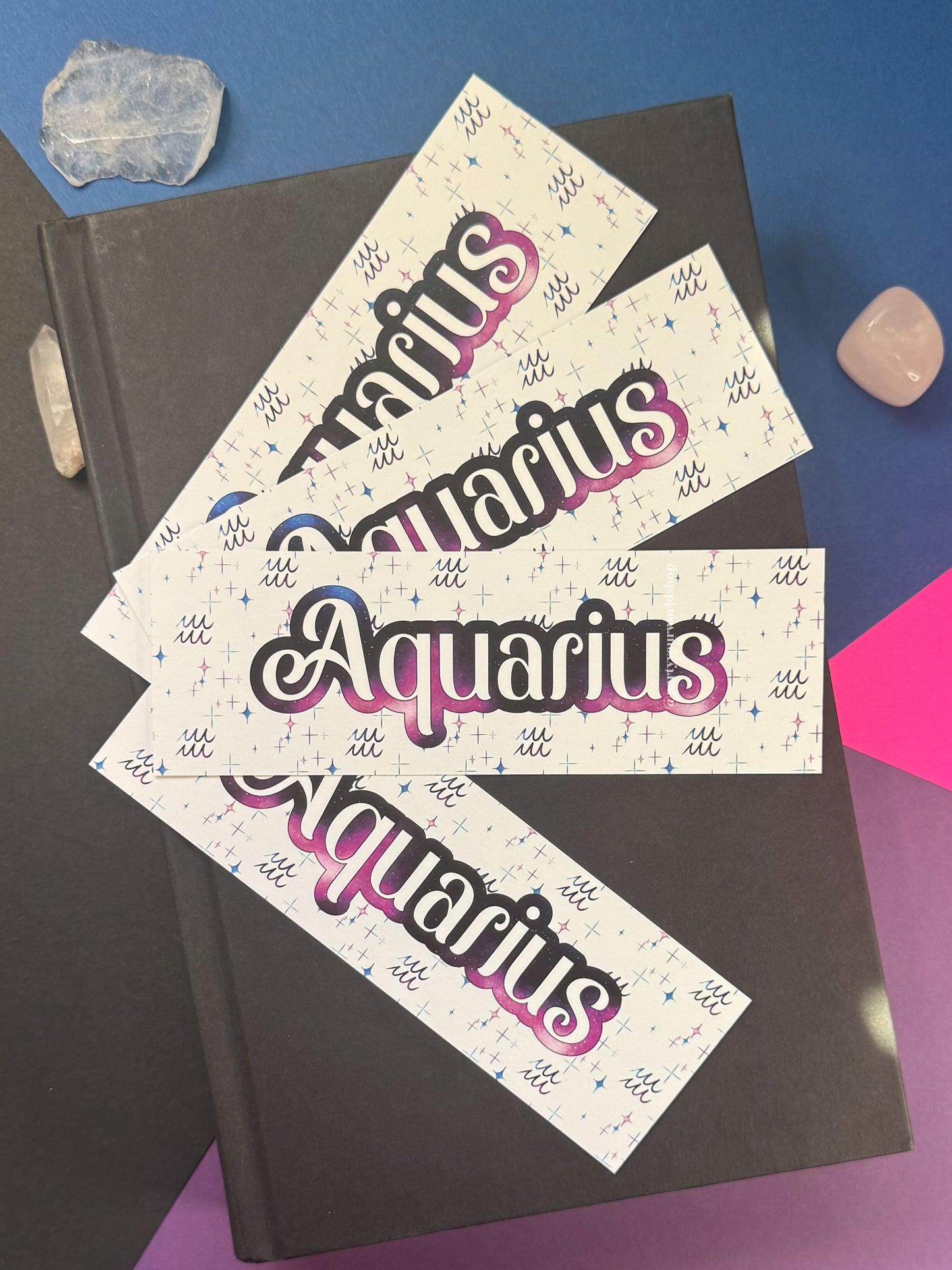 Four rectangular bookmarks with the word "Aquarius" in large stylized lettering, arranged on top of a dark notebook. The tags have a cream background with small blue and pink decorative marks and a pink-to-purple gradient outline around the word.