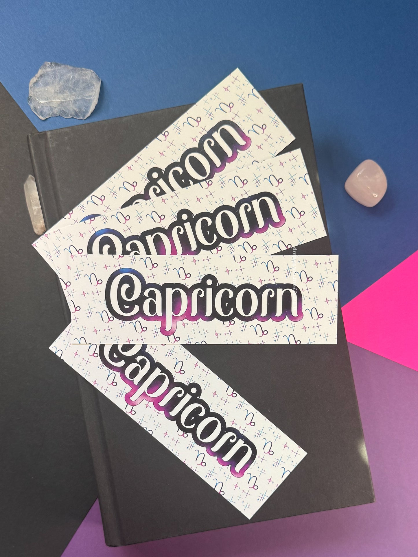 Four white rectangular bookmarks  with the word 'Capricorn' in large black letters outlined in pink and blue, arranged on a black book with crystal stones on a blue and purple background.