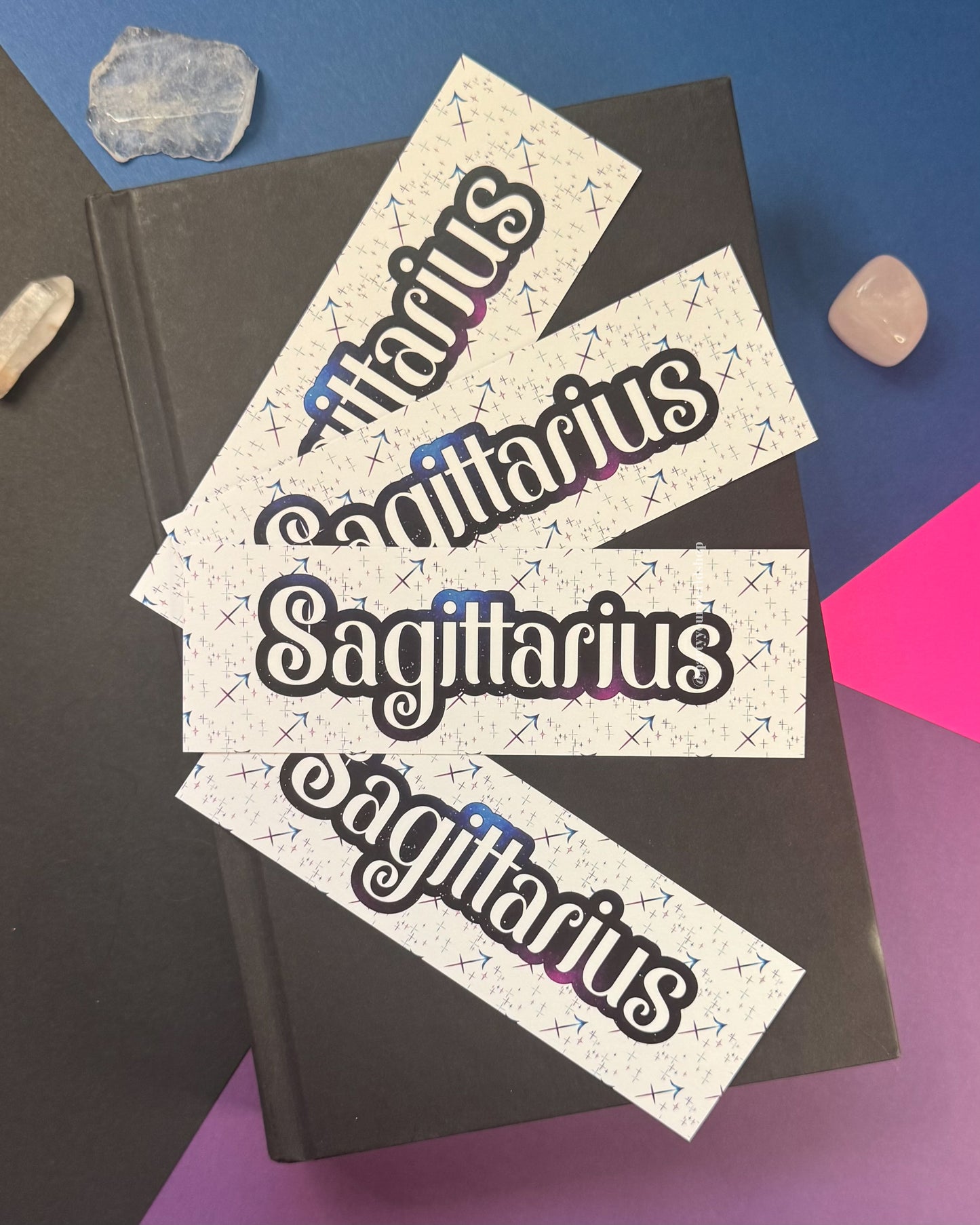 Three white paper bookmarks with the word 'Sagittarius' in a black outlined script and small blue zodiac symbols, arranged on top of a dark book against a colorful blue, pink, and purple background with crystals.