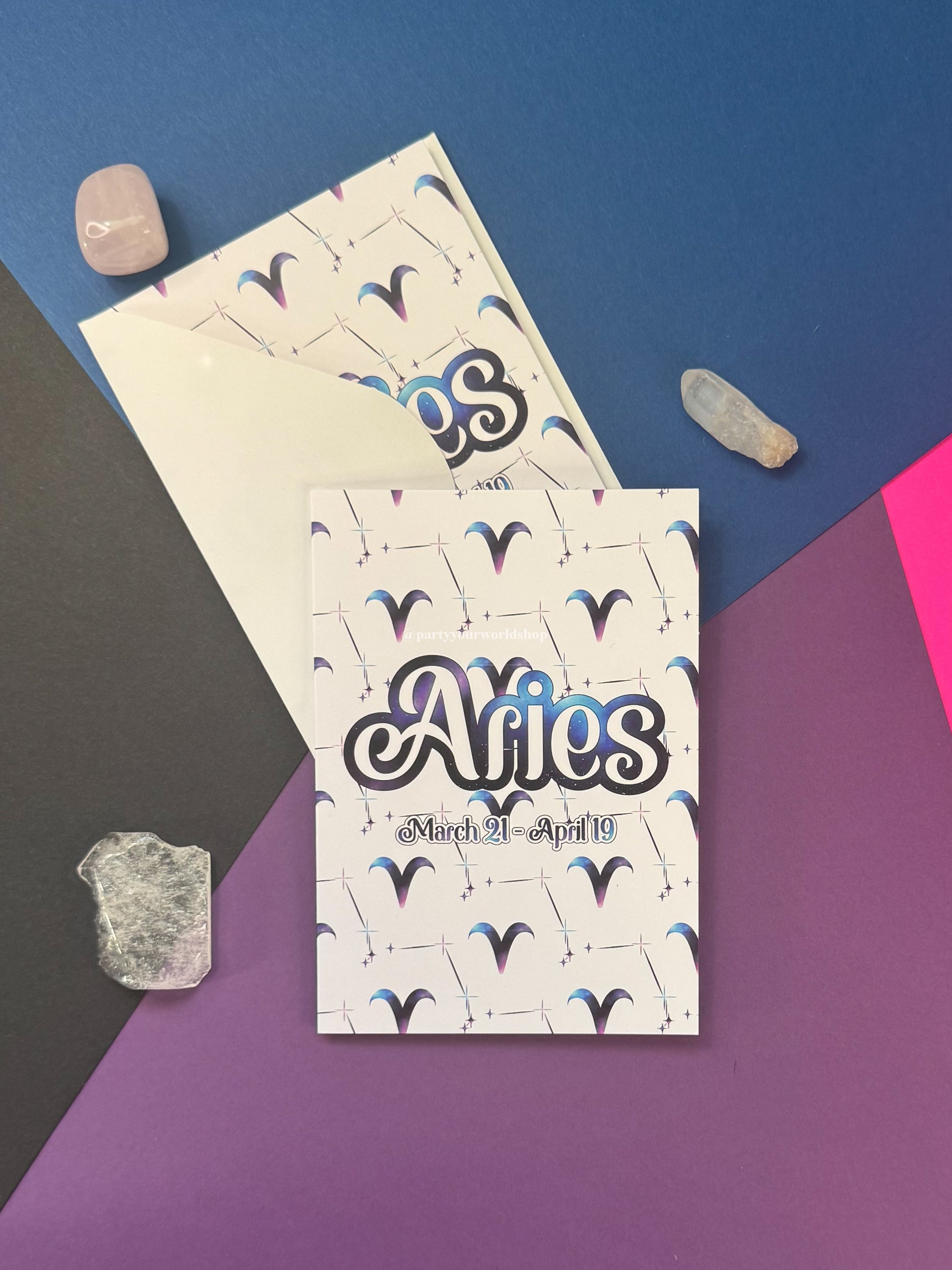 A flat-lay of an Aries zodiac card and matching envelope on a geometric blue, purple, and pink background, styled with small crystals.