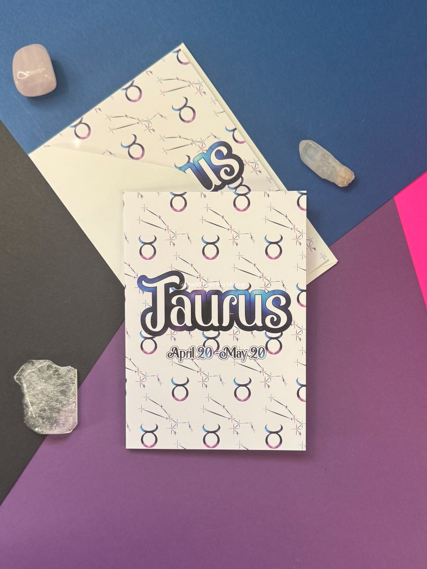A Taurus zodiac-themed greeting card and matching envelope laid flat on a geometric multicolor background with crystal props. The card features the word 'Taurus' in stylized blue and black lettering with the date 'April 20 - May 20' below it, and a repeating zodiac symbol pattern on a white base.