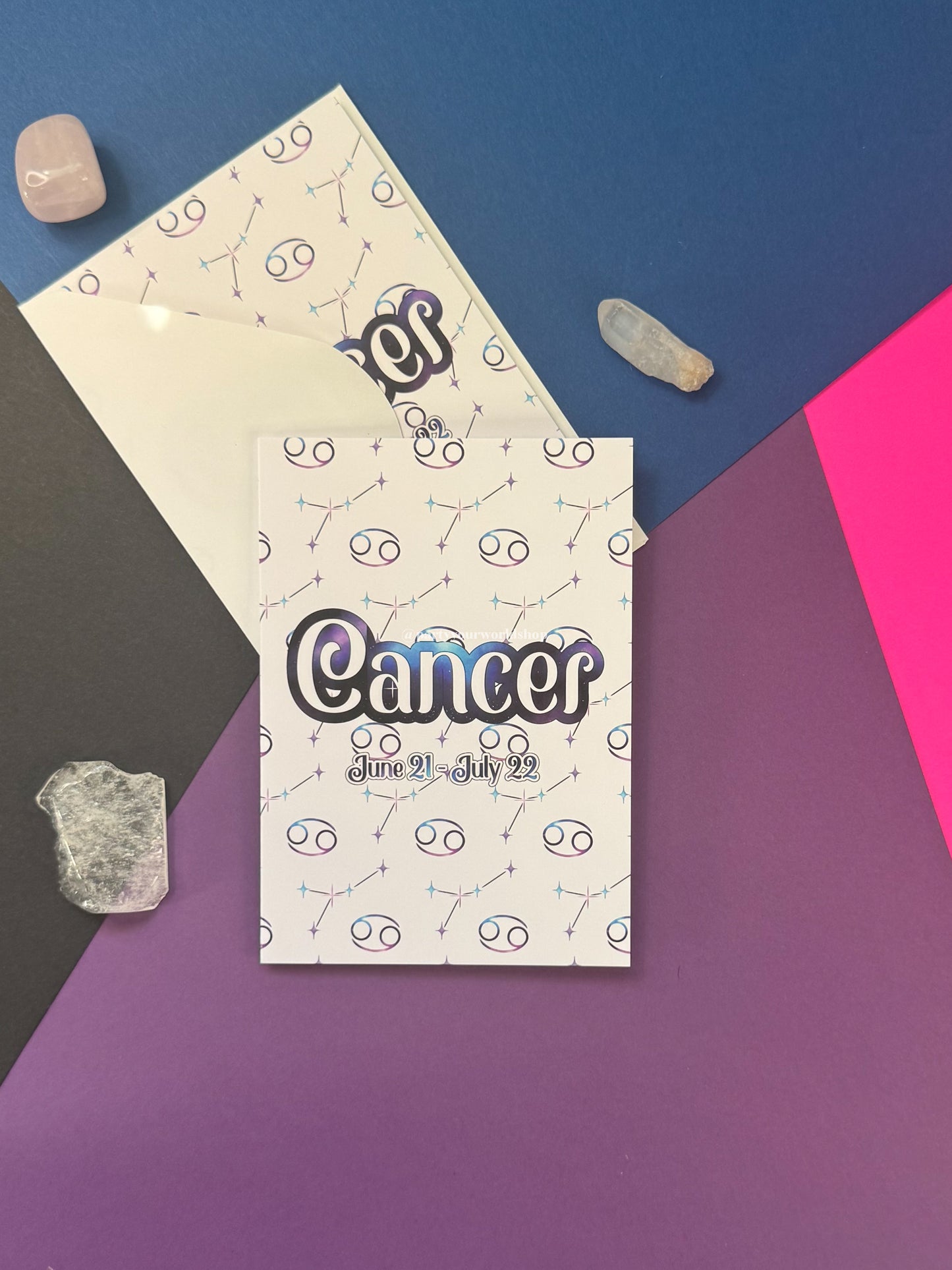 A flat-lay of a Cancer zodiac card and matching envelope on a geometric multicolor background with crystal props. The card features a white background with a repeating pattern of Cancer zodiac symbols and the word 'Cancer' in large black bubble letters with a blue-purple shadow.