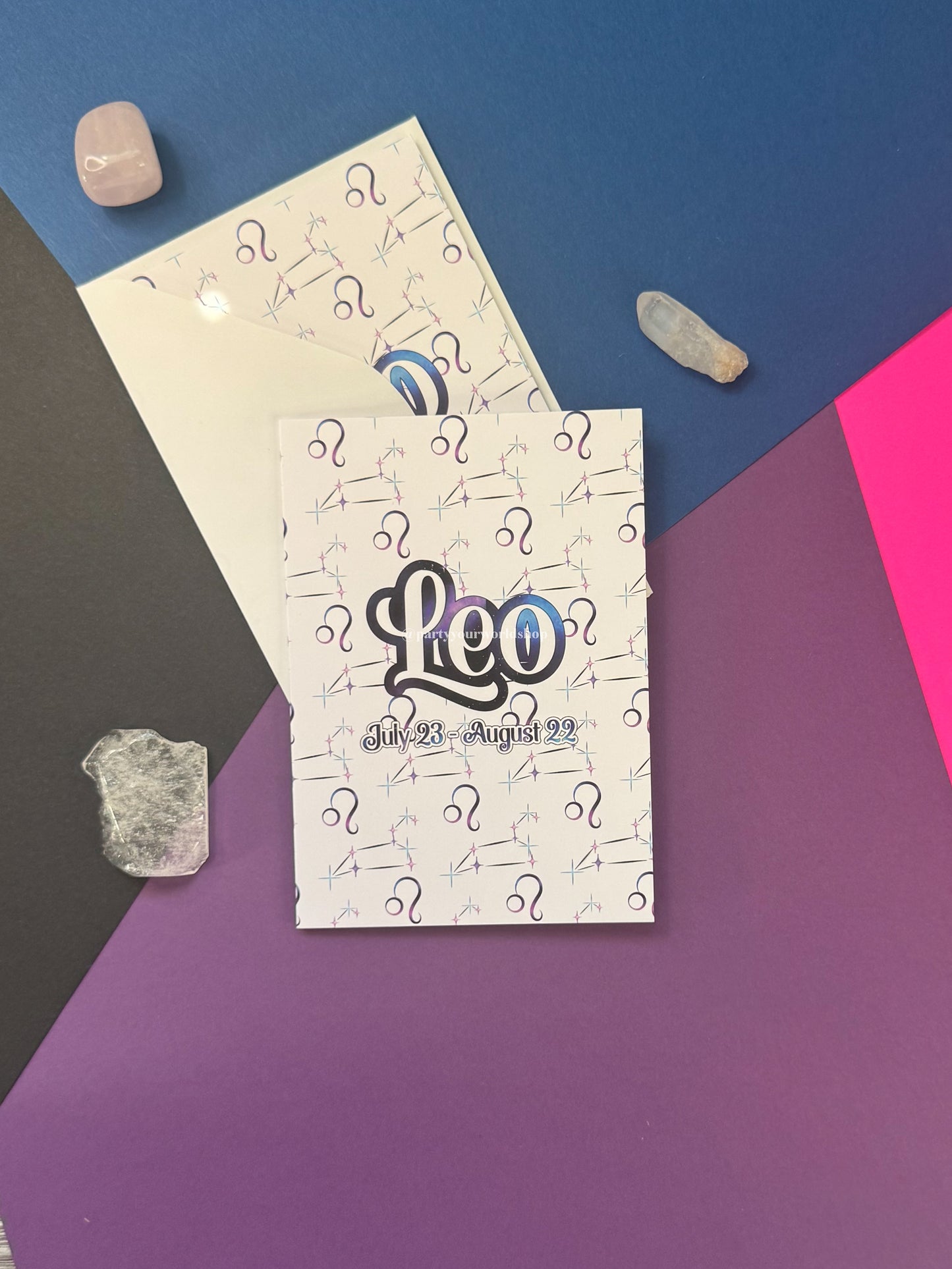 A printed card or booklet titled 'Leo' with the dates 'July 21 - August 22' and repeated question-mark graphics, placed on top of another matching card. The set is styled on a geometric multicolor background with pink, purple, blue, and dark gray sections, plus small crystals.