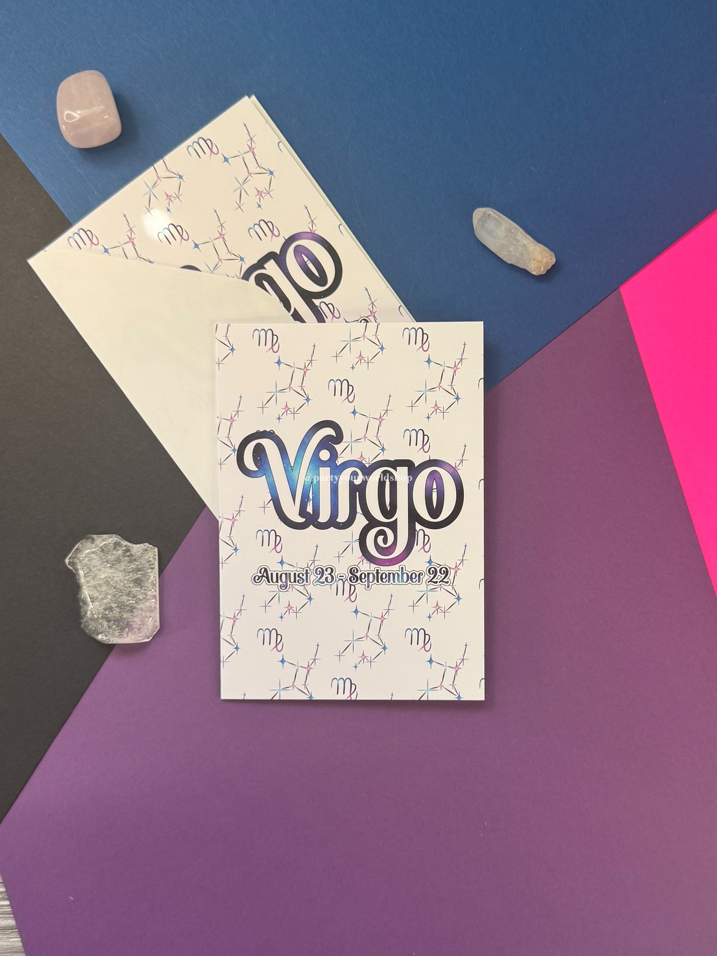 A printed card featuring the word 'Virgo' in large outlined lettering with a purple-to-blue gradient, plus zodiac constellation lines and the date 'August 23, September 22.' The item is photographed on a geometric multicolor background with crystals placed around it.