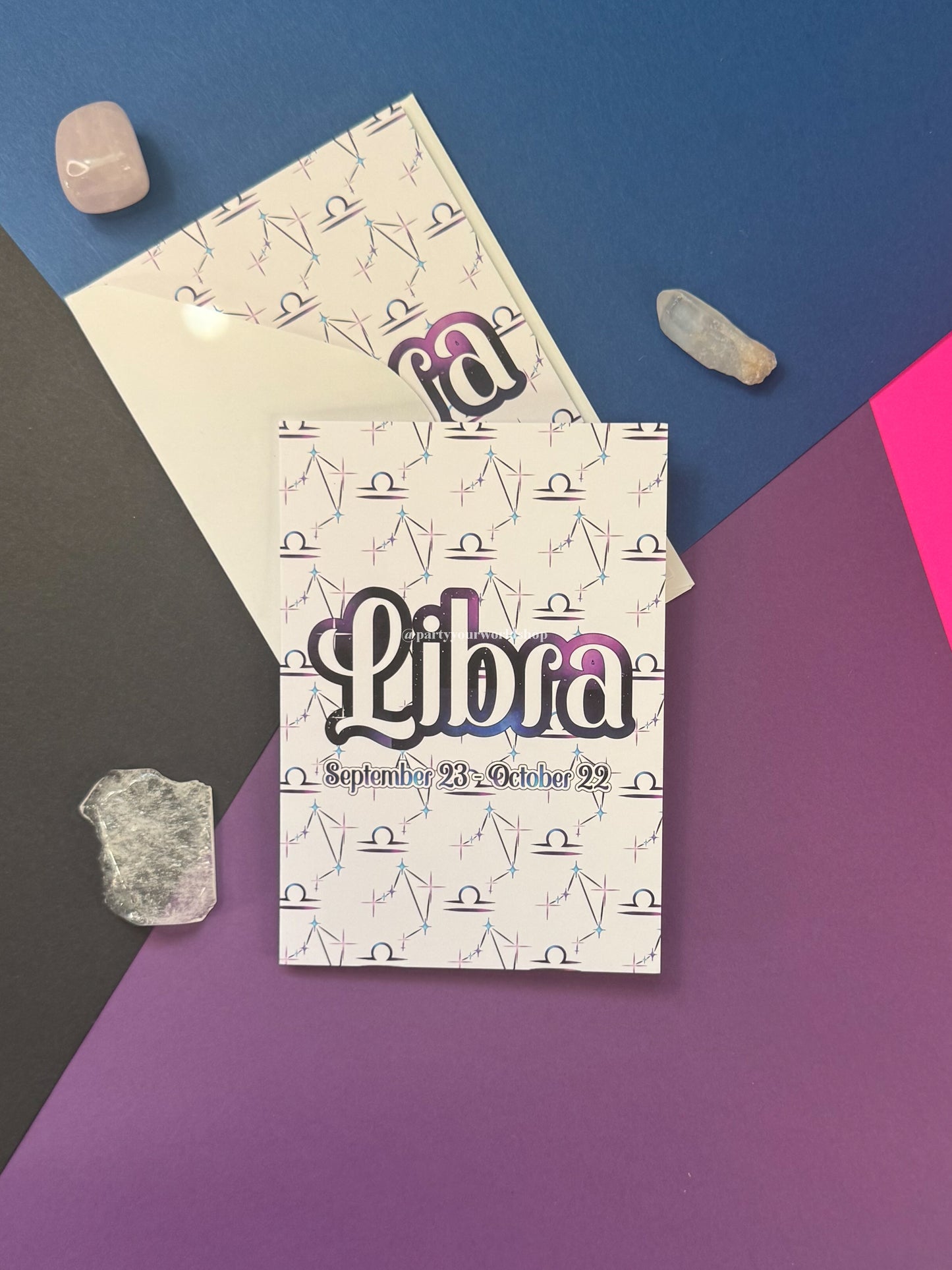 A Libra-themed card  with a white cover patterned with small black line drawings and the word Libra in large stylized lettering, placed on a geometric multicolor background with crystals around it.