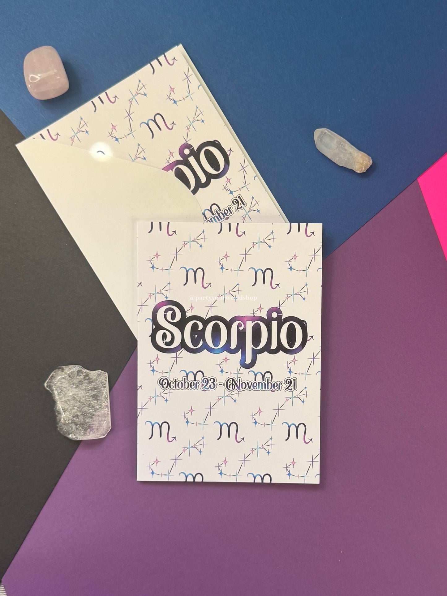 A Scorpio zodiac-themed card  displayed on a geometric blue, purple, and dark gray background with crystal stones around it. The front piece shows the word 'Scoprio' in large stylized lettering with the dates 'October 23 - November 21' below, and a repeating zodiac symbol pattern.