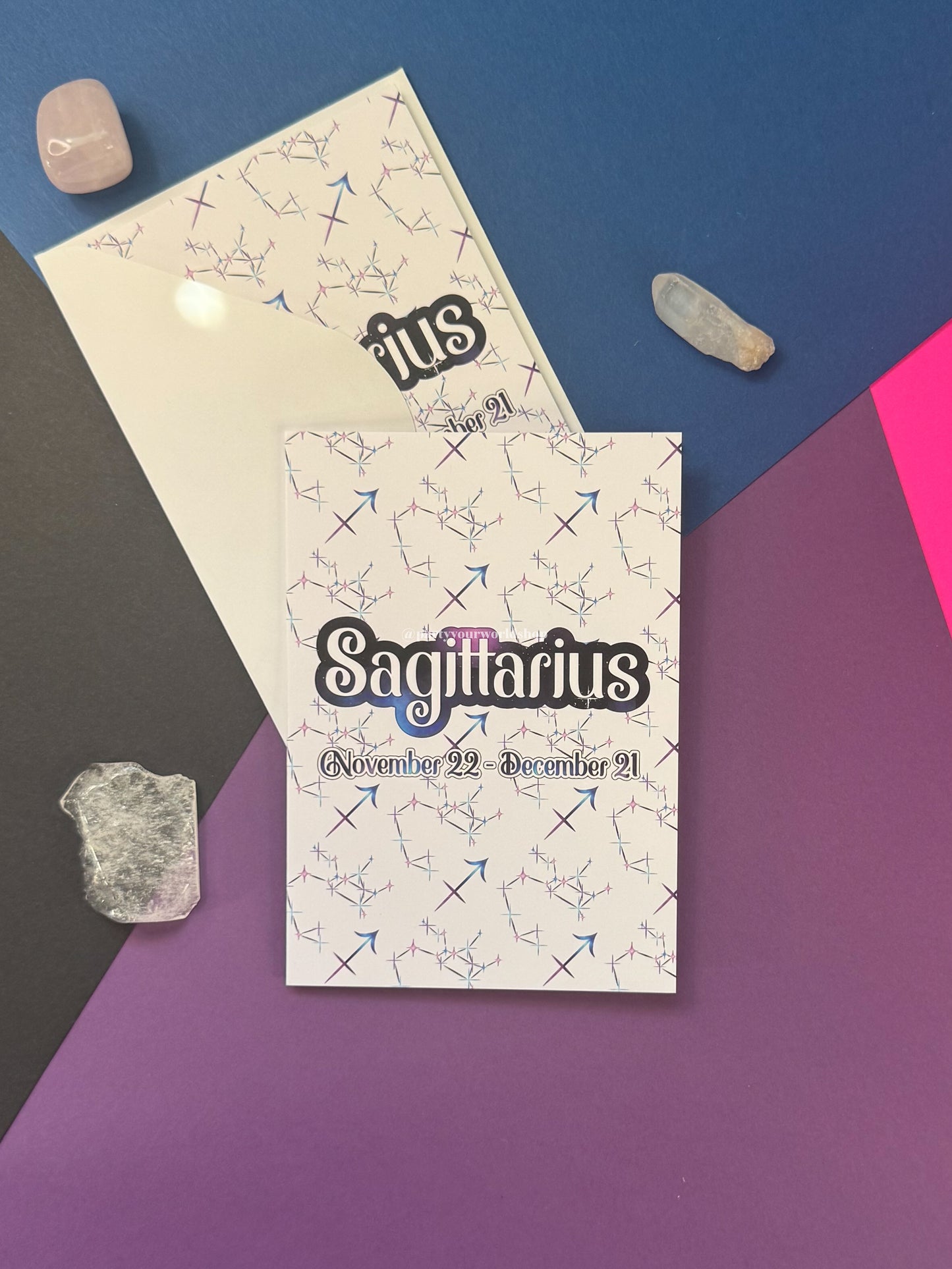 A flat-lay of a white card printed with the word 'Sagittarius' in large black script, surrounded by constellation line drawings and the dates 'November 22 - December 21'. A second cream-colored sheet is partially visible behind it.