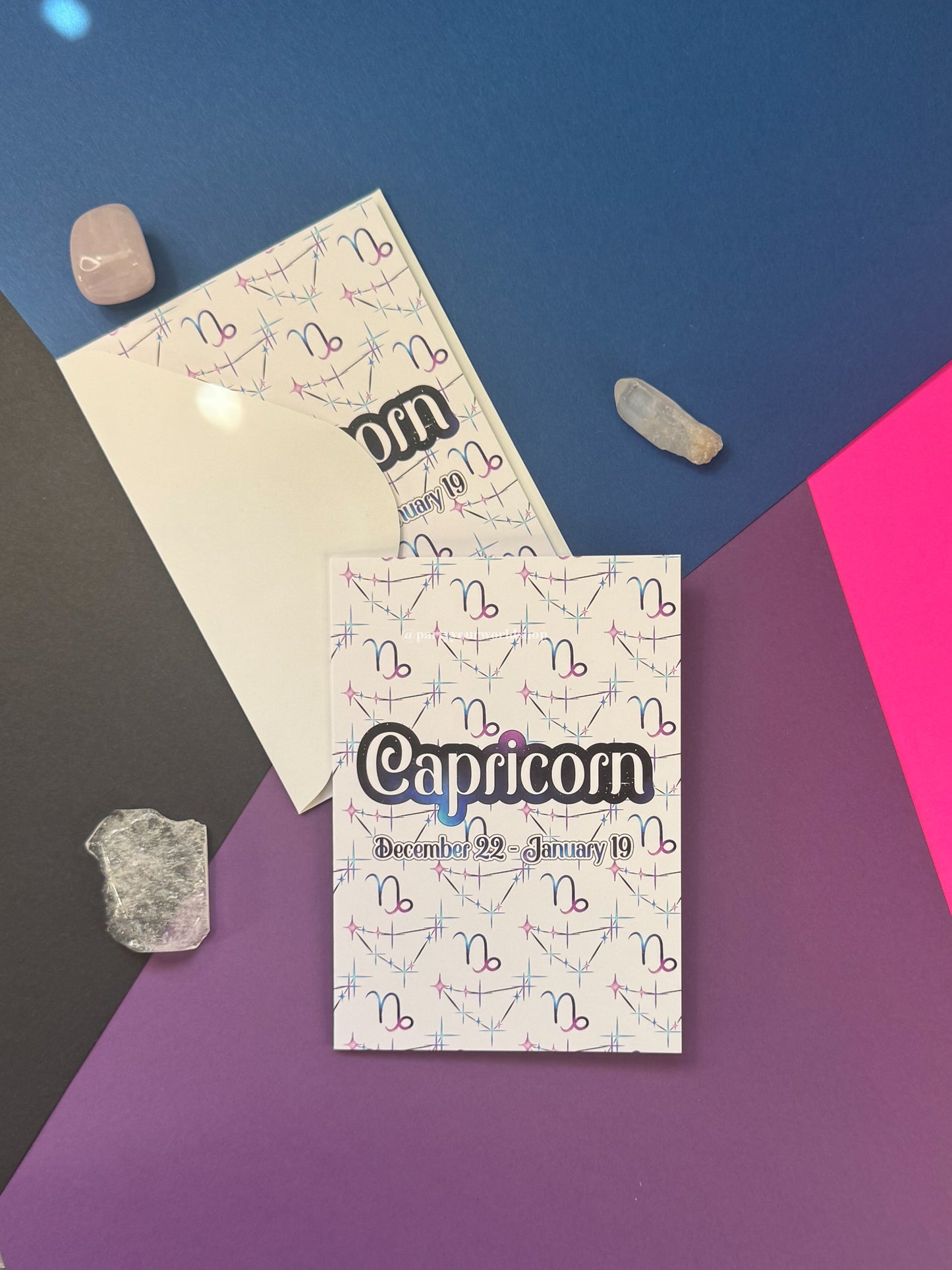 A printed Capricorn card with a repeating astrological symbol pattern, placed on top of a cream envelope on a geometric multicolor background with crystals.
