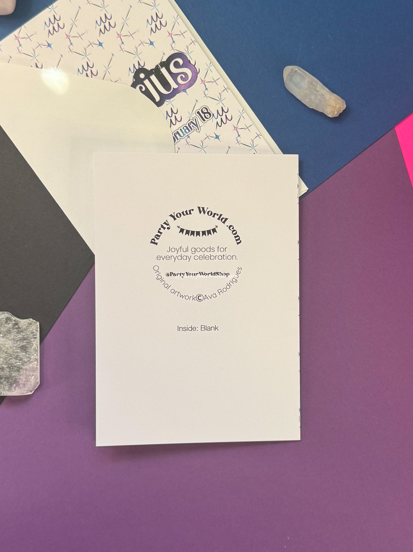 A white greeting card or insert with black printed text reading 'Party Your World.com' and 'Inside: Blank' lies on a purple surface, with part of a patterned white envelope or paper visible behind it and colorful flat-lay props around it.