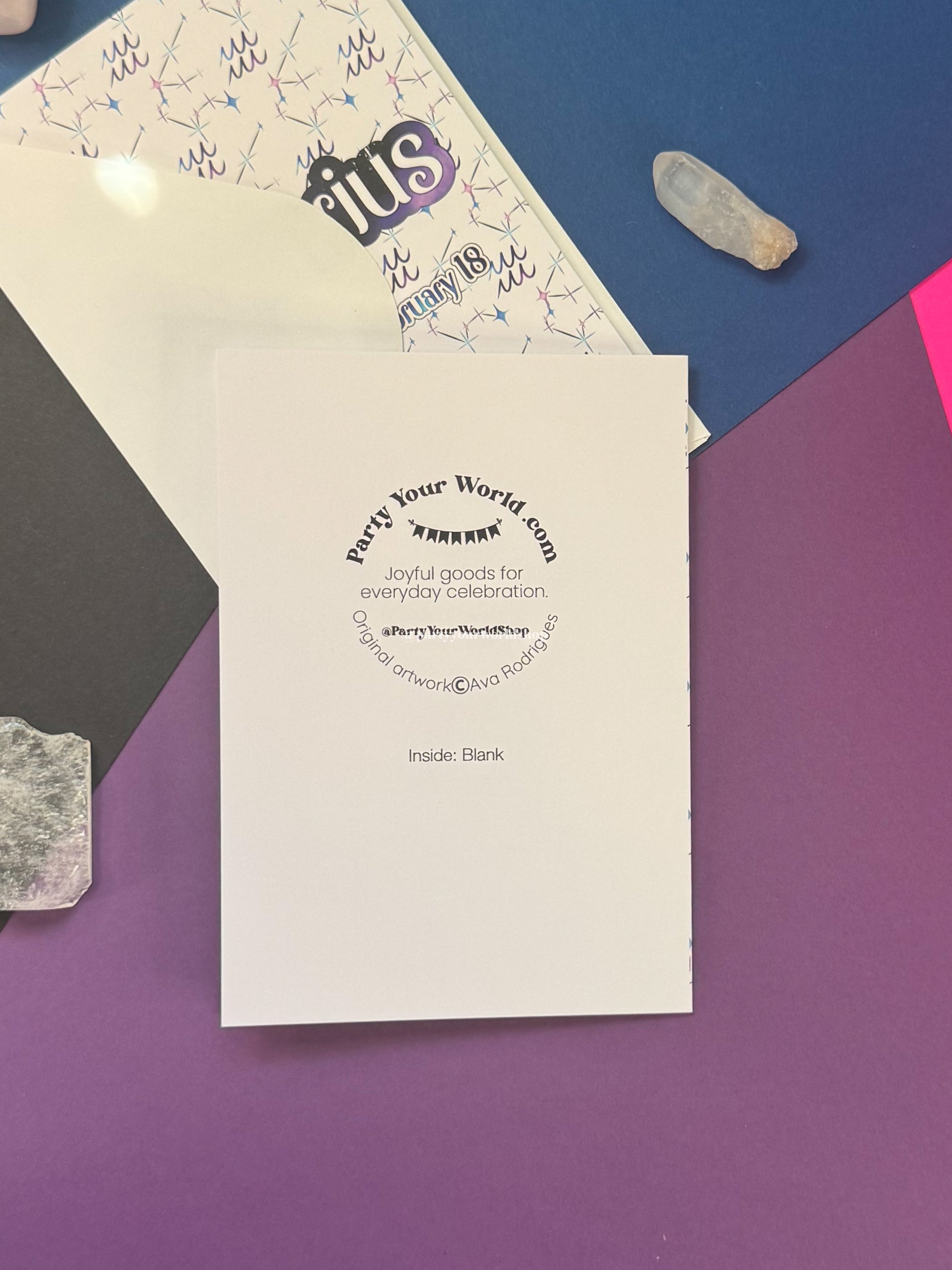 A white greeting card or insert with black printed text reading 'Party Your World.com' and 'Inside: Blank' lies on a purple surface, with part of a patterned white envelope or paper visible behind it and colorful flat-lay props around it.
