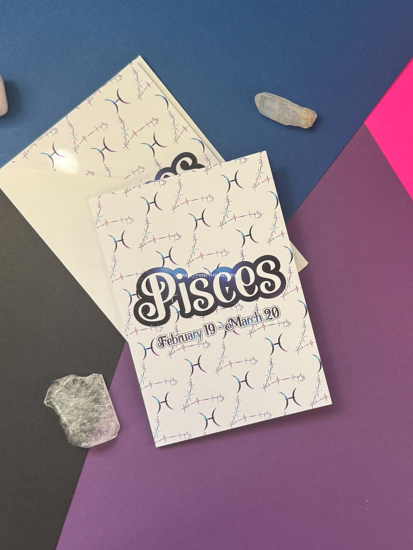 A Pisces zodiac card set featuring a white card with black and blue astrological symbols and the word 'Pisces' in large script, shown with a cream envelope on a geometric blue, purple, pink, and gray background with small crystal props.