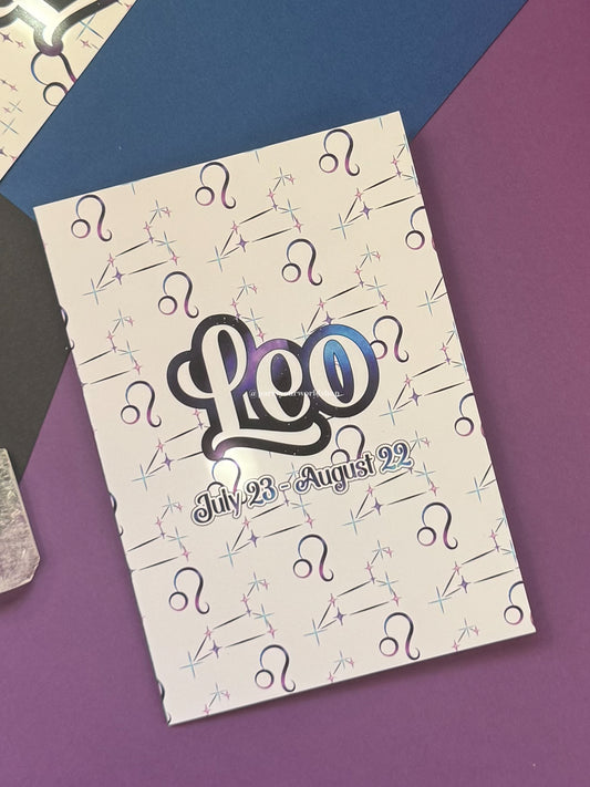 A square white card featuring the word 'Leo' in large stylized black and blue-purple lettering, with 'July 23 - August 22' below it. The cover is covered in a repeating pattern of astrological Libra zodiac symbols and is photographed on a purple surface with a blue geometric shape in the background.